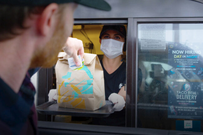 Taco Bell worker wearing mask at drive-thru next to hiring sign